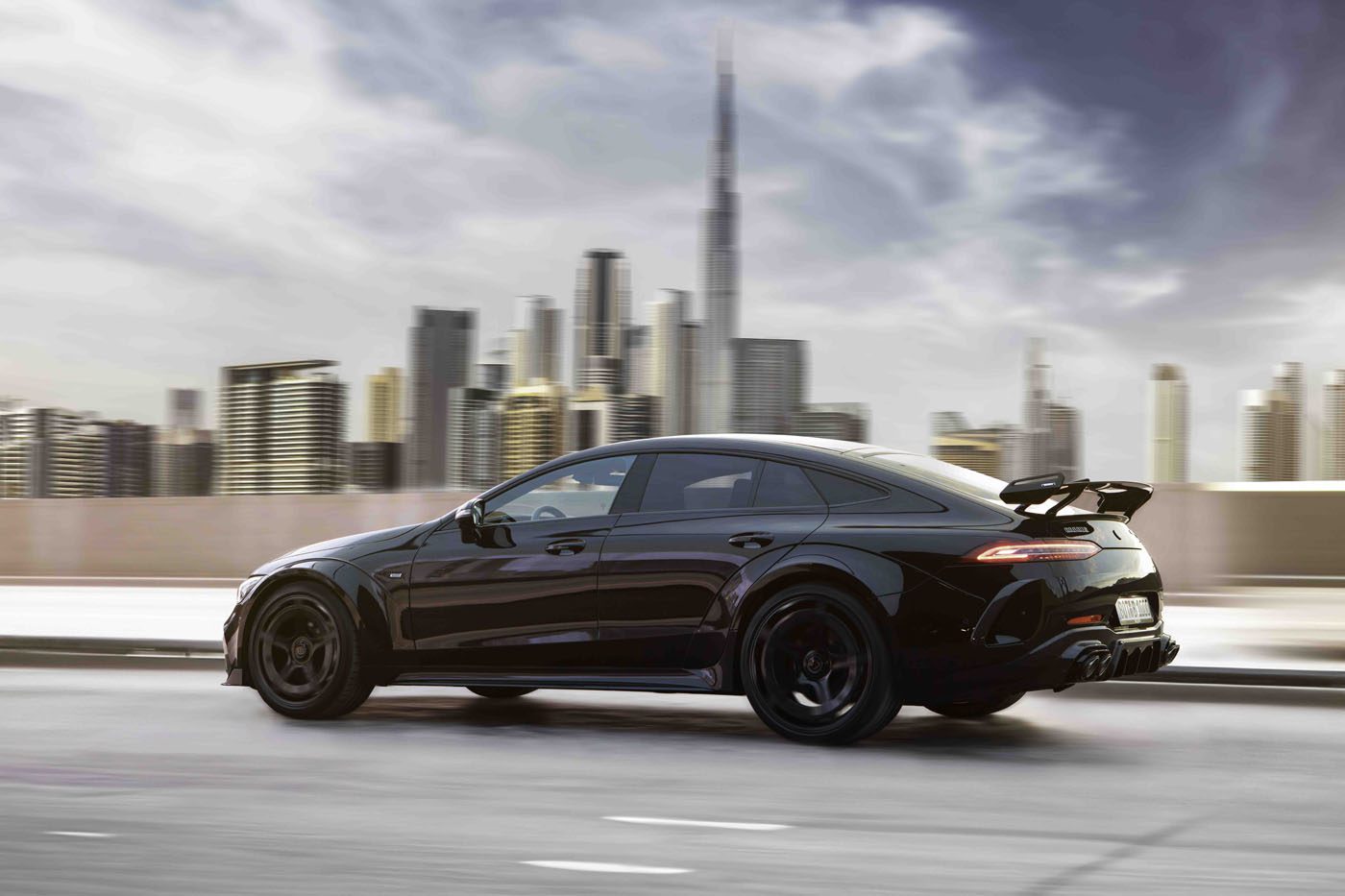 A BRABUS Rocket 1000, a luxury car, speeds along a city road with towering skyscrapers and a tall tower in the background beneath a cloudy sky.