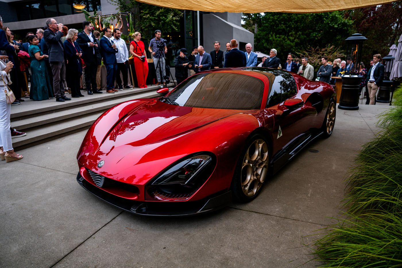 A red Alfa Romeo 33 Stradale supercar is displayed outdoors at an event, surrounded by a crowd of people on concrete steps and under a canopy.