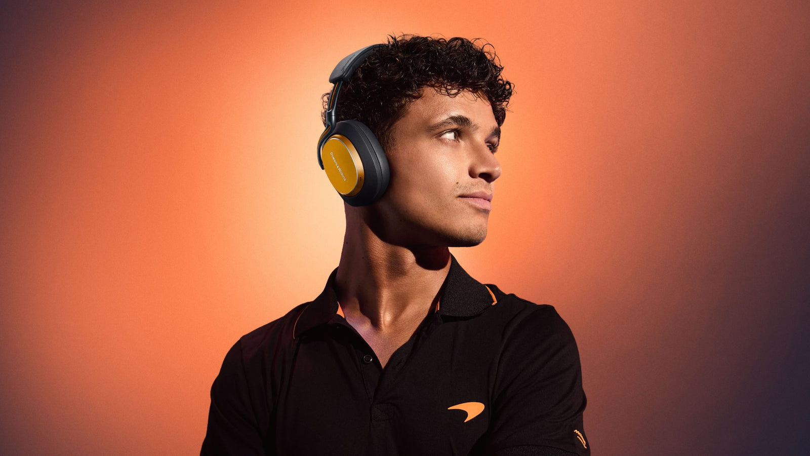 A young man wearing large over-ear Bowers & Wilkins Px8 S2 headphones and a black polo shirt with an orange logo stands against an orange gradient background.