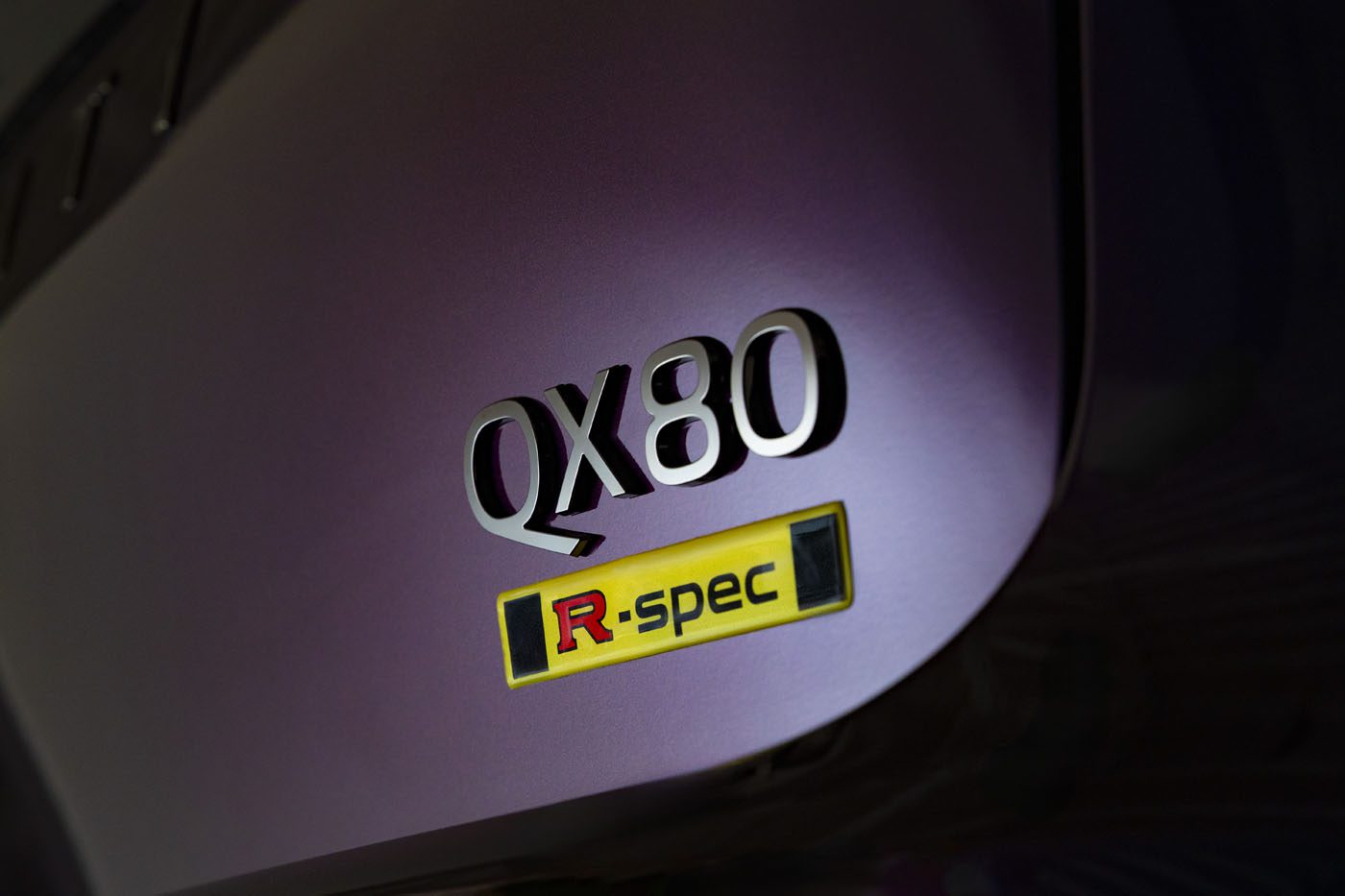 Close-up of a vehicle badge showing "QX80" and a yellow "R-spec" emblem on a polished, purple-painted surface—this striking Infiniti is a GT-R-powered QX80 showcased at SEMA.