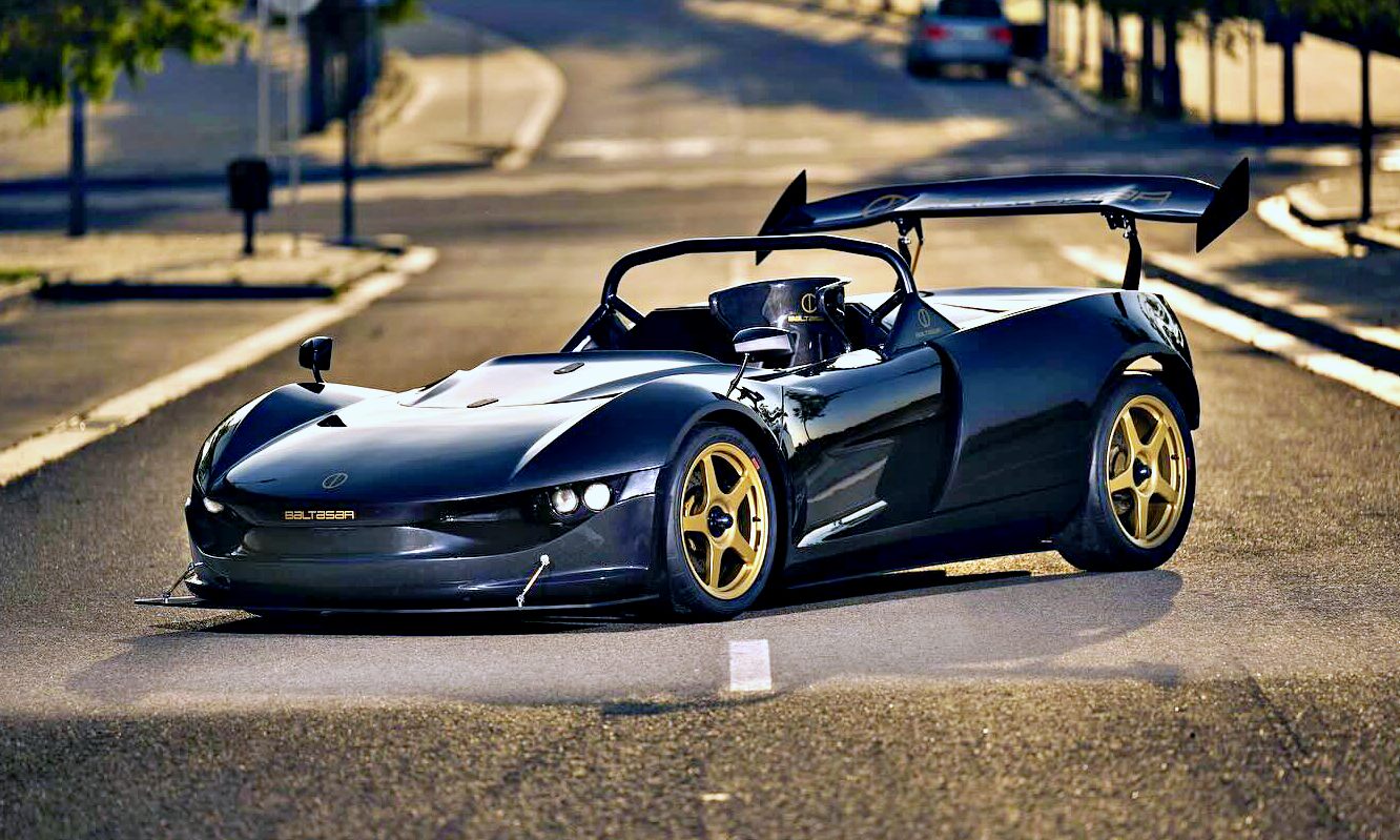 A black open-top sports car with gold magnesium wheels, an FIA cage, and a large DRS wing is parked on a sunlit street—a new king EV of track days?.