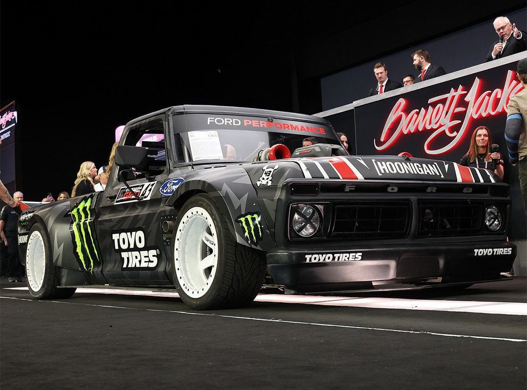 Black custom race truck with Ford branding, white wheels, and sponsors' logos including Toyo Tires and Monster Energy, showcased indoors at the Scottsdale Auction by Barrett-Jackson, with people in the background.