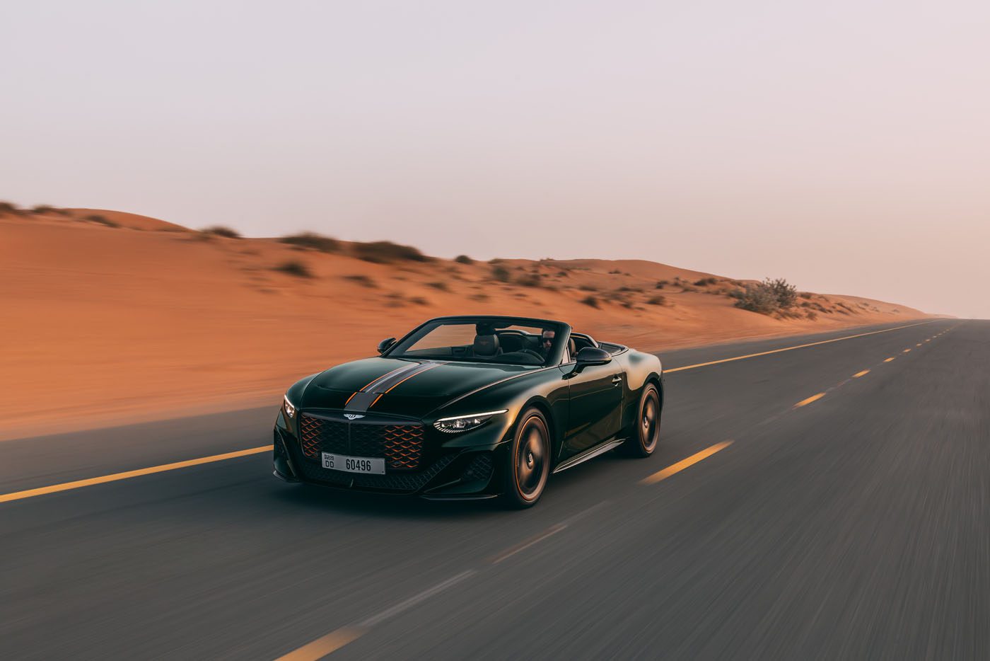 A dark green Bentley Batur Convertible makes its public debut, driving on an empty road through a desert landscape at sunset near Dubai.