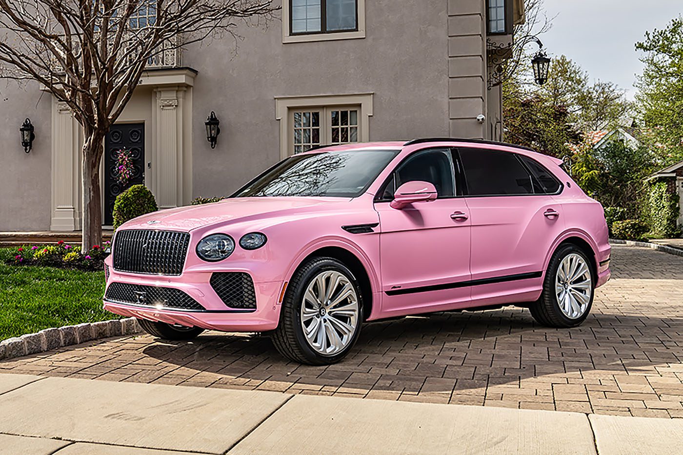 A Pink Bentayga SUV is parked on a brick driveway in front of a beige house with landscaped greenery, showing off one of the unique Bentley exterior colors.
