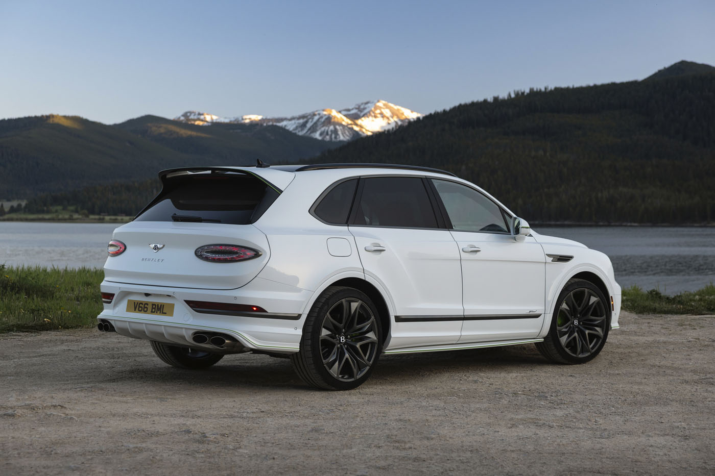 A powerful Bentayga, this white luxury SUV is parked on a dirt road by a lake, with mountains and trees in the background under a clear sky.