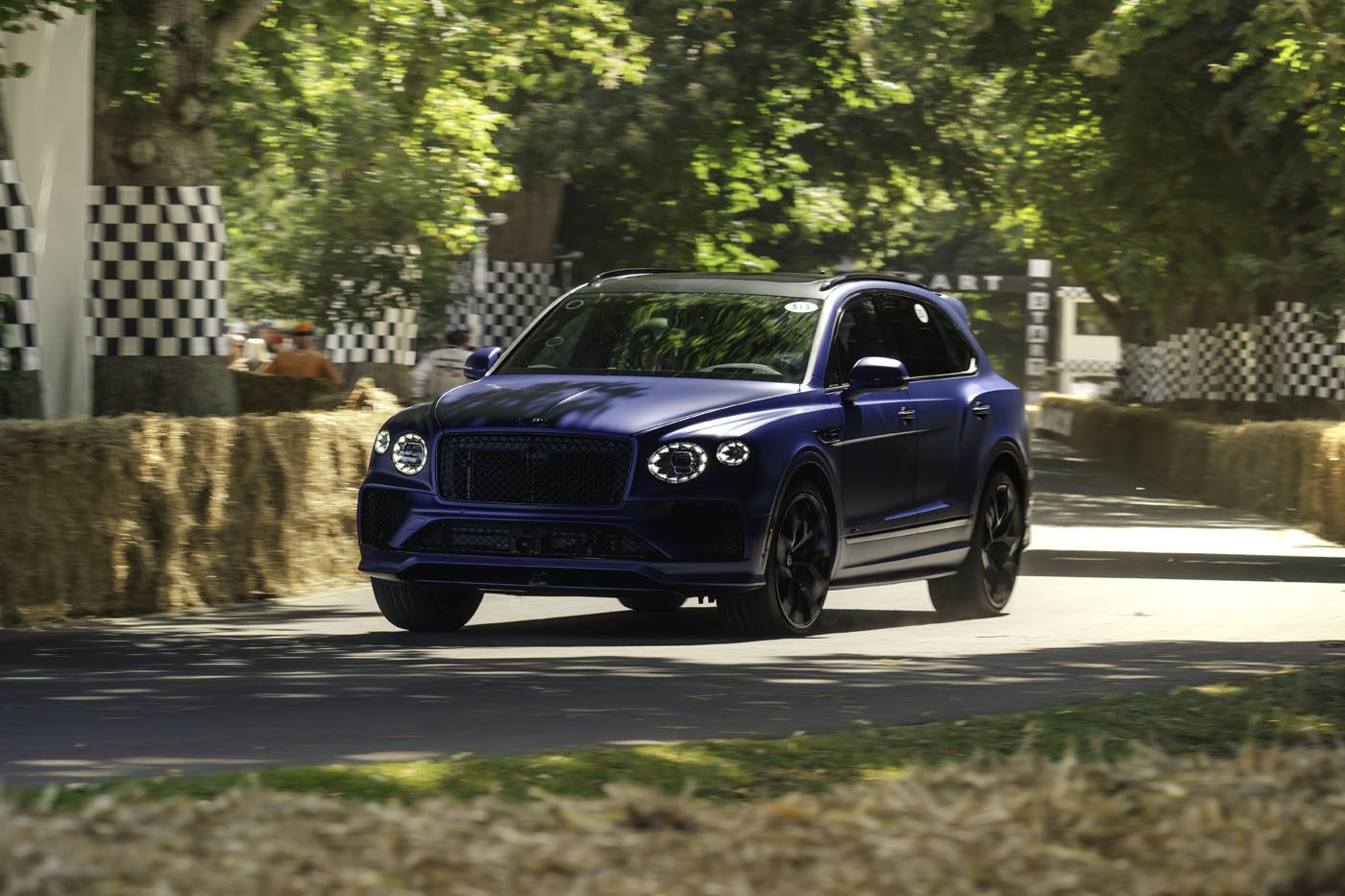 A blue Bentley Bentayga Speed luxury SUV drives on a paved road lined with hay bales and checkered barriers, surrounded by green trees in daylight, reminiscent of the famous Goodwood Hillclimb record runs.