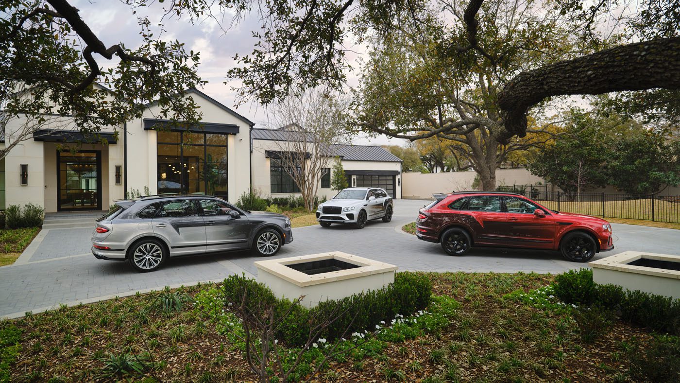 Three luxury SUVs, including bespoke Victoris Bentaygas in silver, white, and red, are parked on a paved driveway. They stand in front of a modern house with large windows, surrounded by trees and lush landscaping.