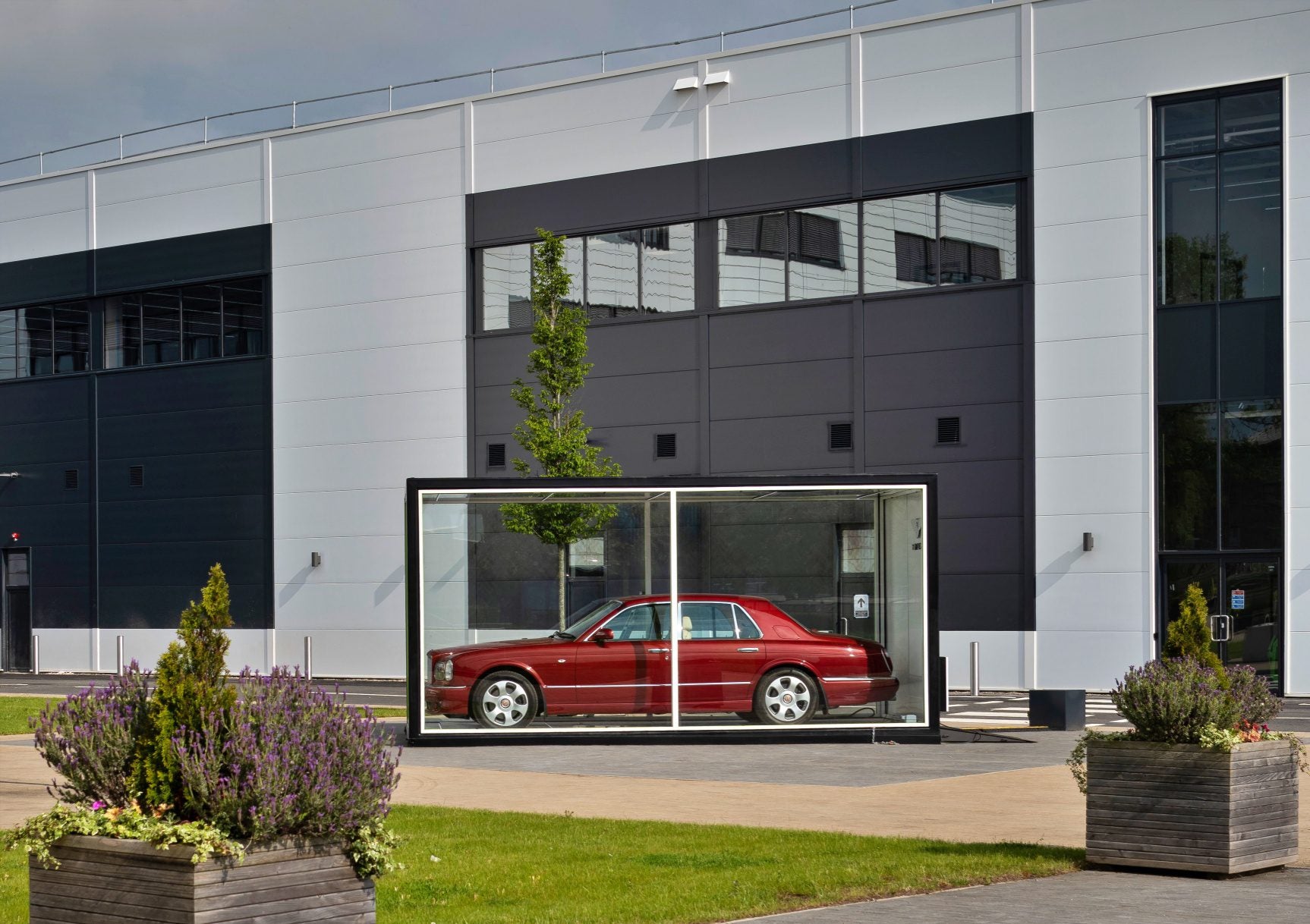 A red Bentley is displayed inside a transparent glass case outside a modern Tech Center, symbolizing the brand's move toward electrification against the building's gray and white exterior walls.