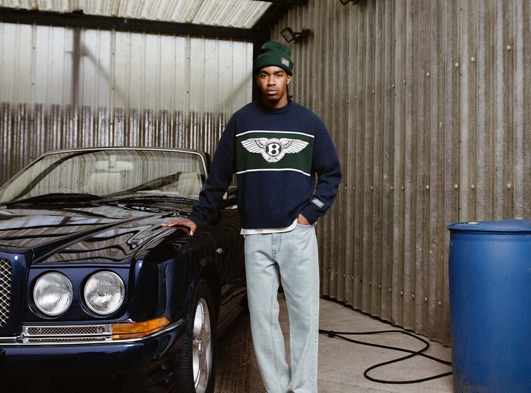 A person in a beanie and a Capsule Collection Bentley sweater stands beside a blue convertible car in a corrugated metal garage.