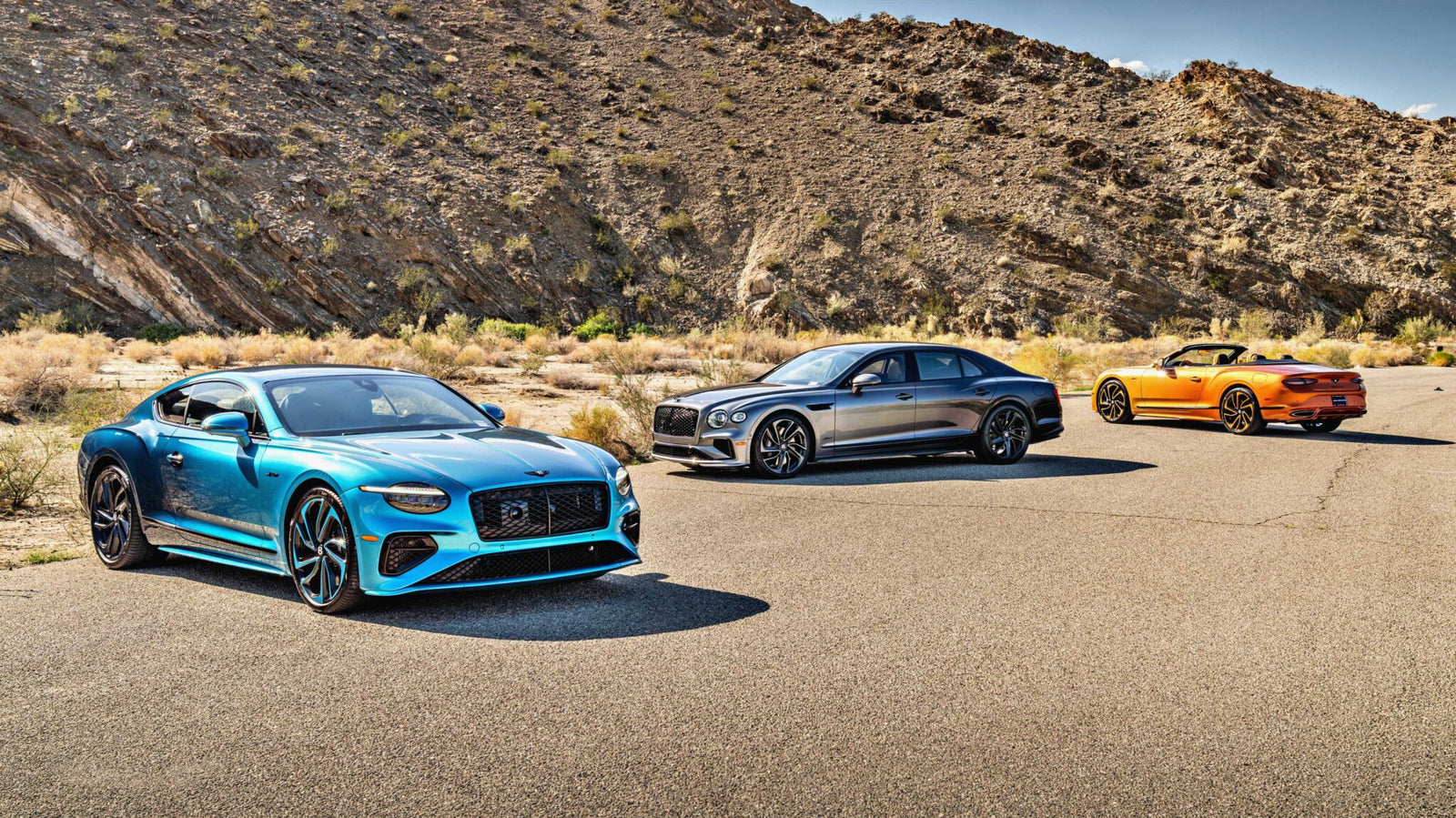 Three luxury cars—a blue coupe, a silver sedan, and an orange convertible—stand on a paved desert road as the First Ombré by Mulliner Collection arrives at Bentley Rancho Mirage with three bespoke commissions.
