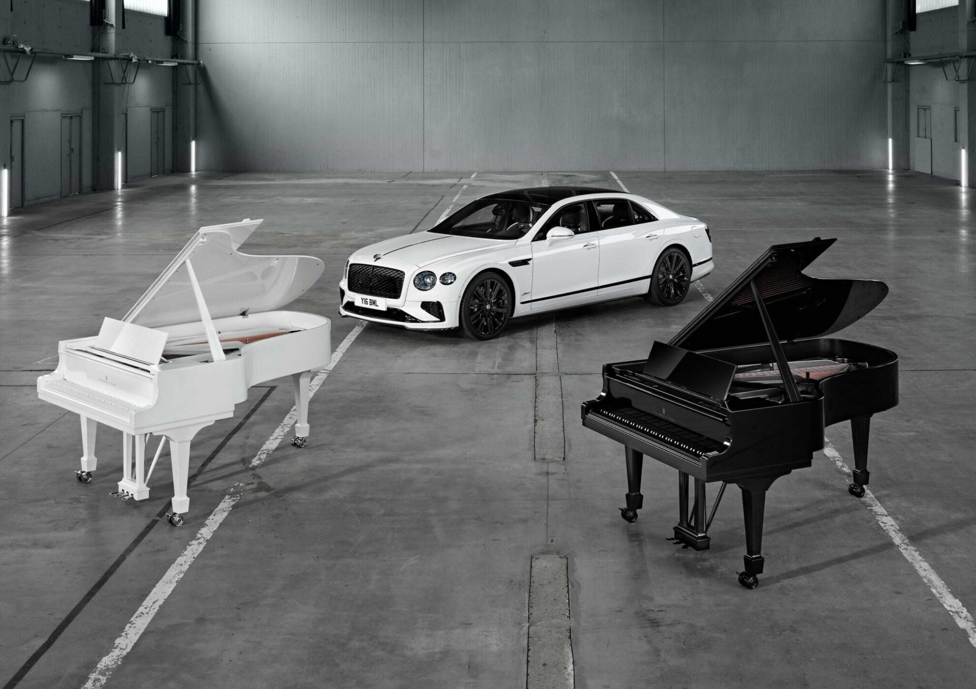 A white luxury car is parked in an empty warehouse between a white grand piano and a black grand piano, as Bentley and Steinway & Sons unite in Hamburg for a rare celebration of craftsmanship.