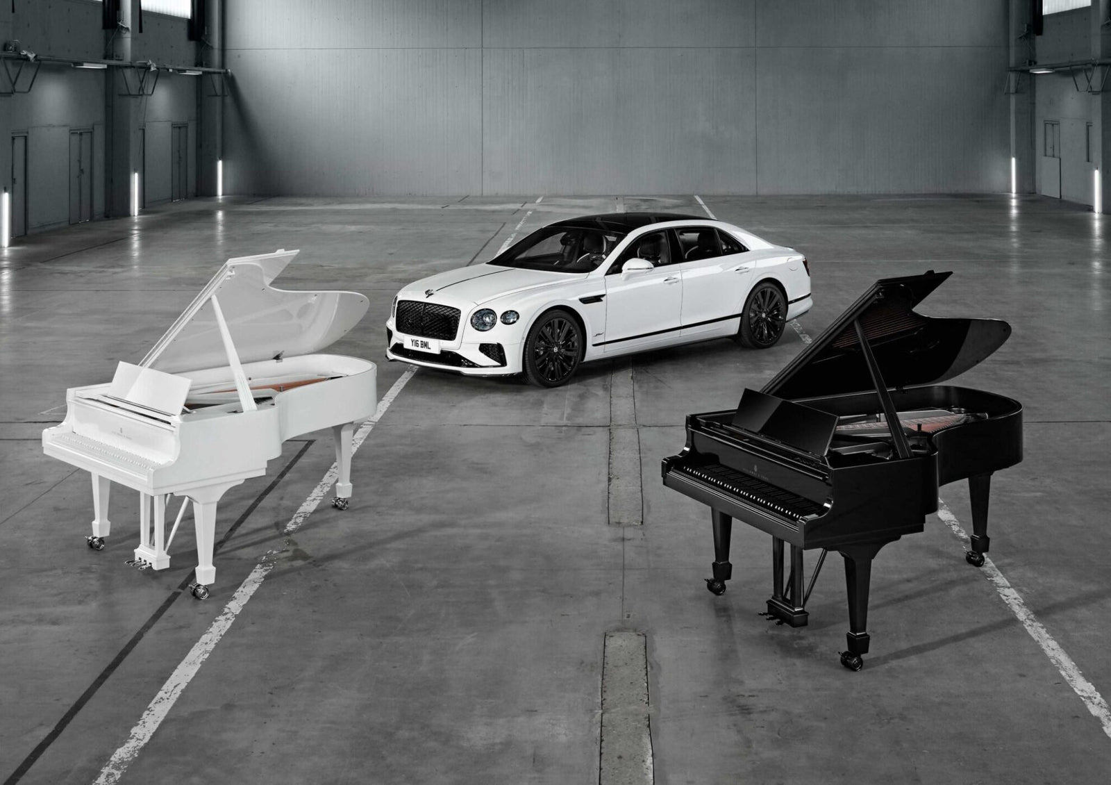 A white luxury car is parked in an empty warehouse between a white grand piano and a black grand piano, as Bentley and Steinway & Sons unite in Hamburg for a rare celebration of craftsmanship.