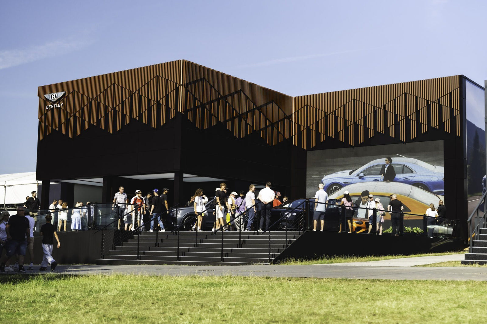 A group of people stand outside a modern Bentley Motors showroom at Goodwood, with a large screen displaying cars and a suited man, on a sunny day.