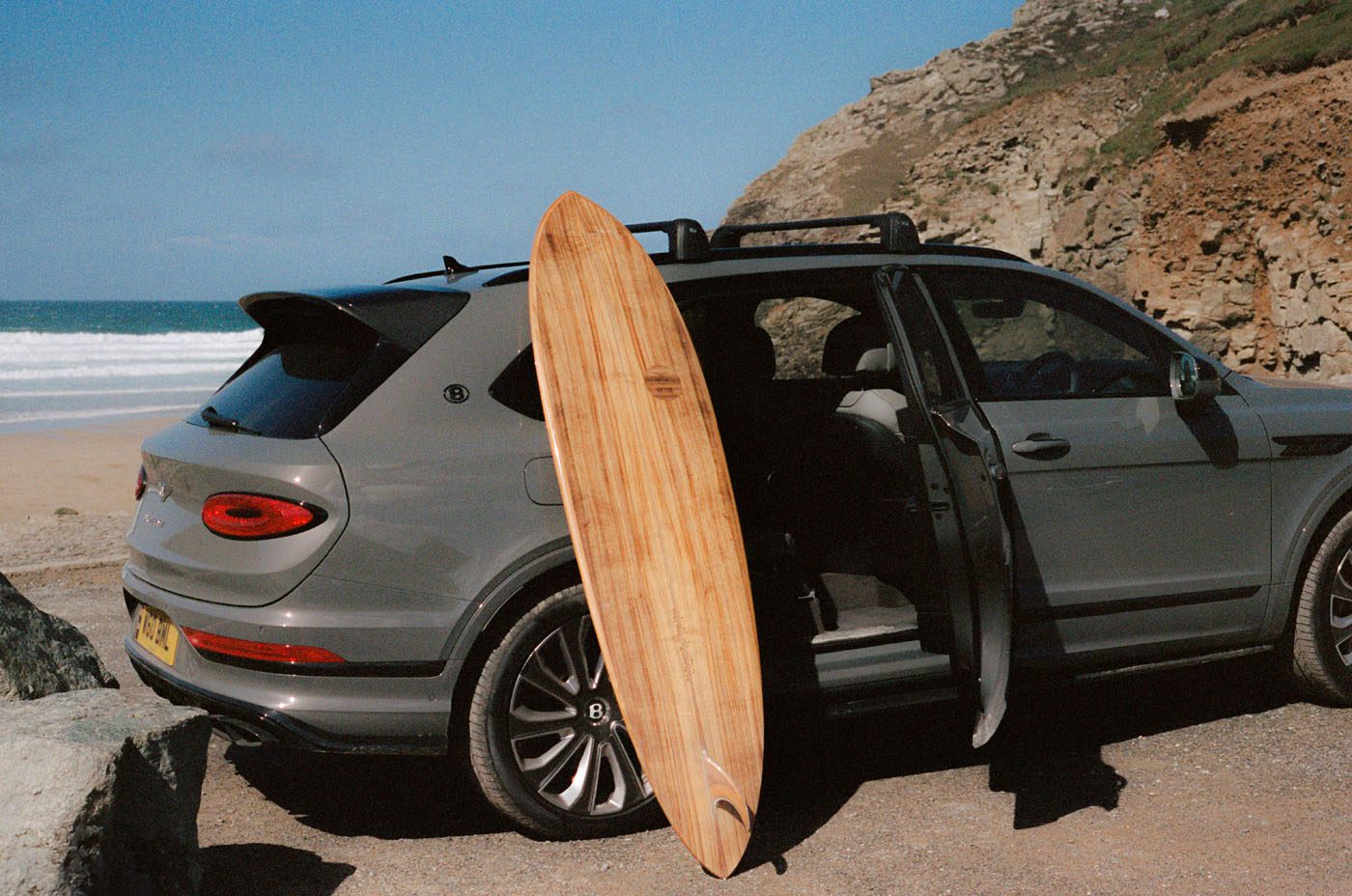 A wooden Otter Surfboards surfboard is propped against a grey SUV parked near a rocky beach with open car doors; the ocean and cliffs are visible in the background, hinting at an inspiring surfboard collaboration.