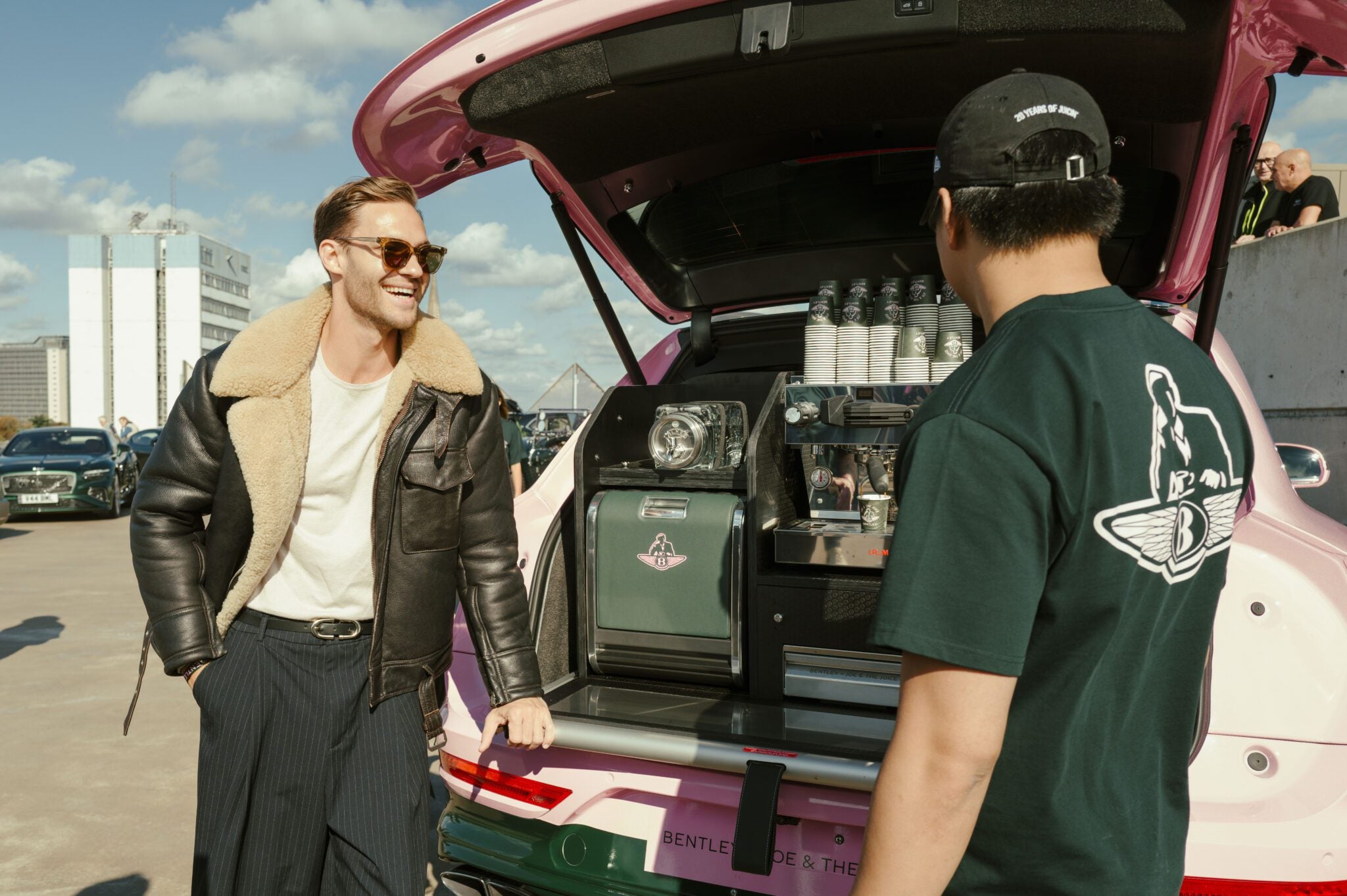 Two men stand by an open Bentley Bentayga trunk, transformed into a stylish Bentayga Coffee Bar with cups and equipment visible, on a sunny day in an urban parking lot.