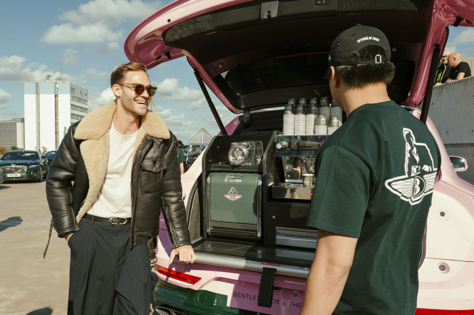 Two men stand by an open Bentley Bentayga trunk, transformed into a stylish Bentayga Coffee Bar with cups and equipment visible, on a sunny day in an urban parking lot.