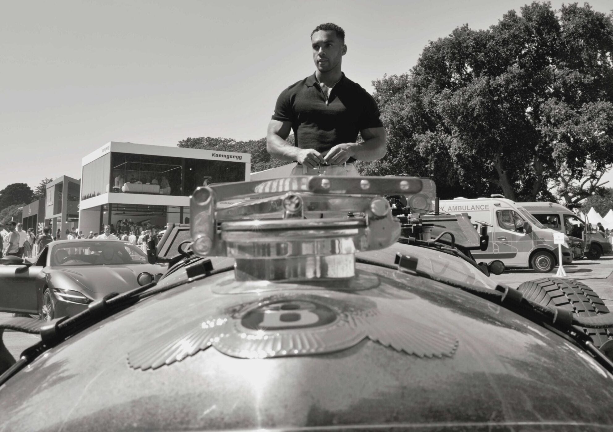 Lucien Laviscount, Global Ambassador, stands behind a vintage Bentley at an outdoor event, with an ambulance and modern buildings in the background.