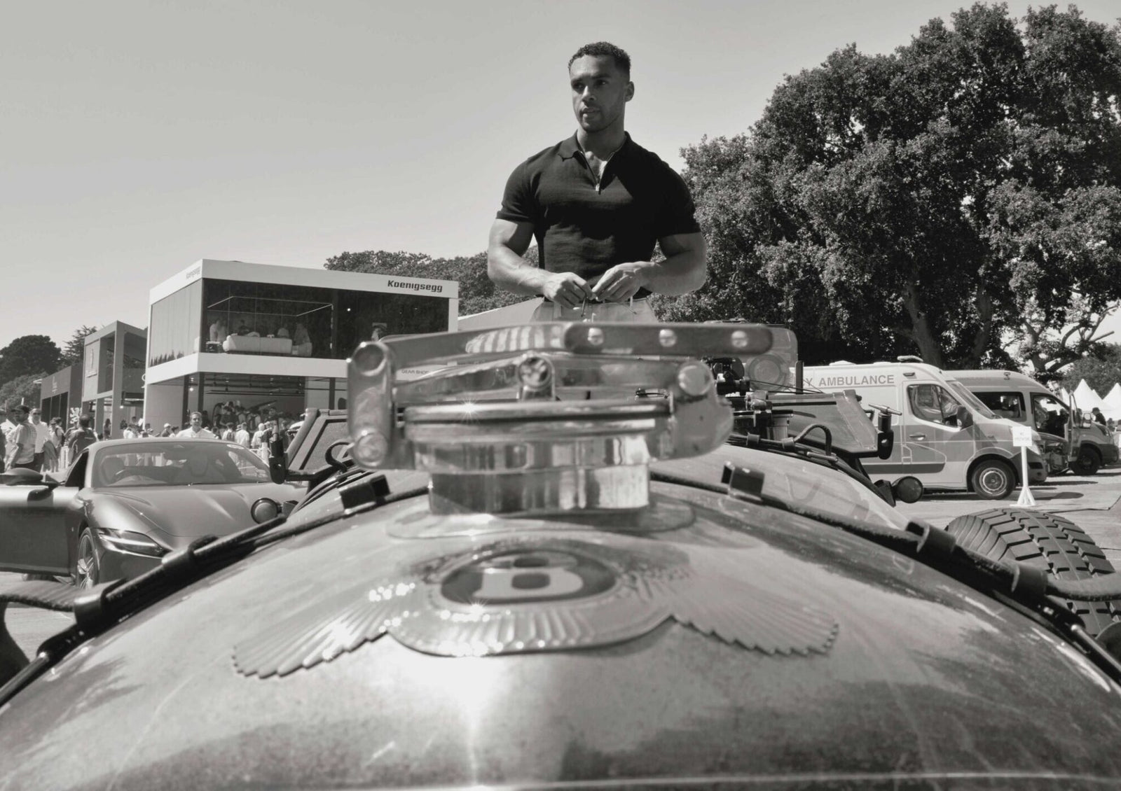 Lucien Laviscount, Global Ambassador, stands behind a vintage Bentley at an outdoor event, with an ambulance and modern buildings in the background.