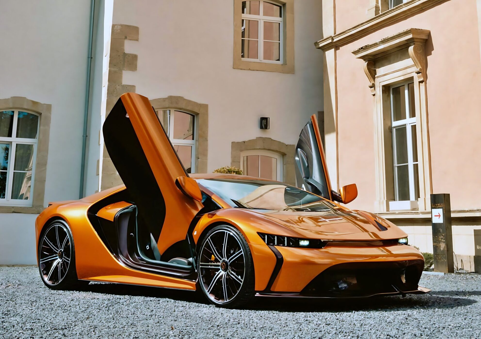 An orange Bertone GB110 meets Arancio Zero—a color that defined an era—with its scissor doors open, parked on a gravel surface in front of a beige building with large windows.