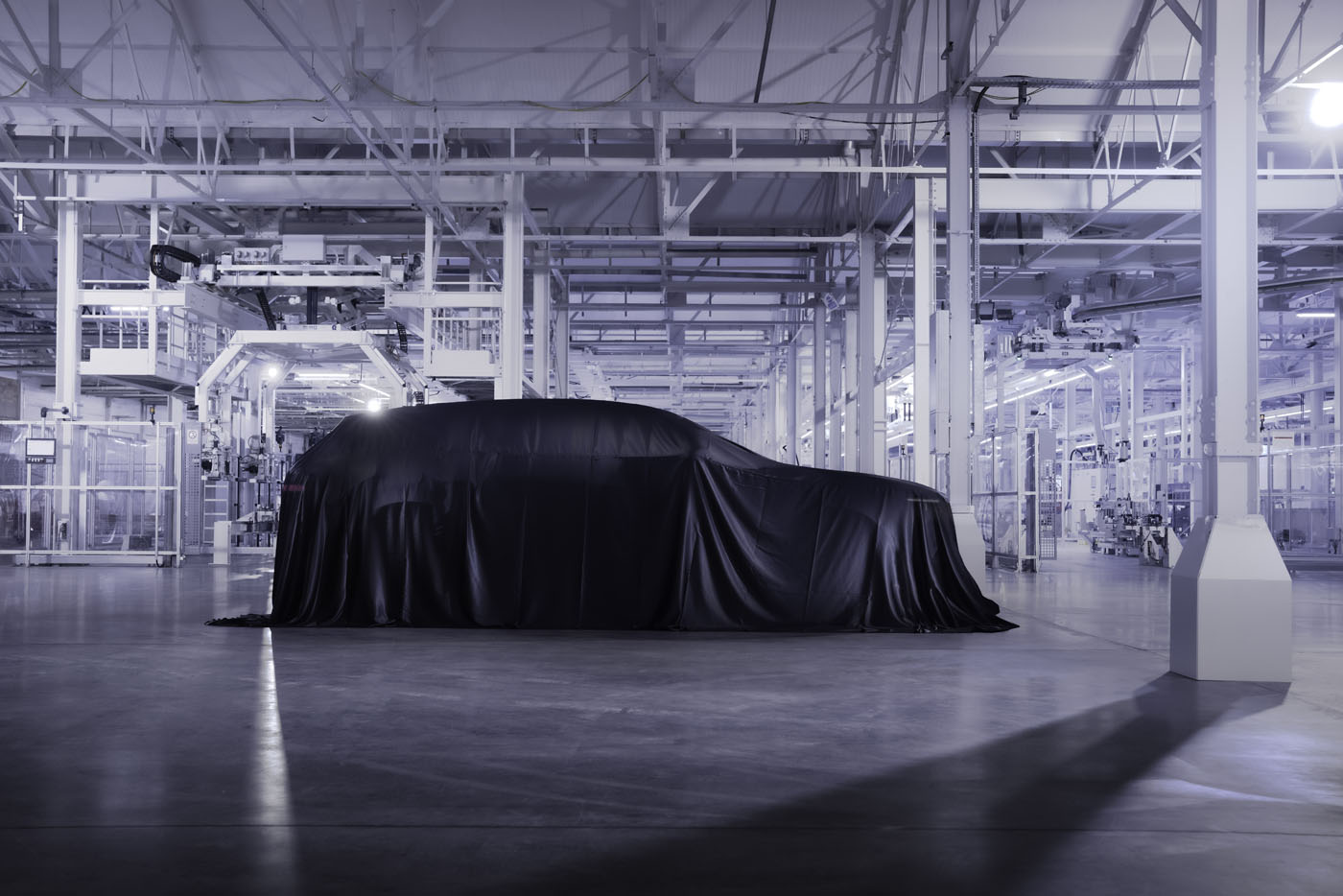 A covered car sits in the center of a modern, brightly lit automotive factory with advanced machinery in the background, hinting at the future of Bentley hybrids and electric vehicles.