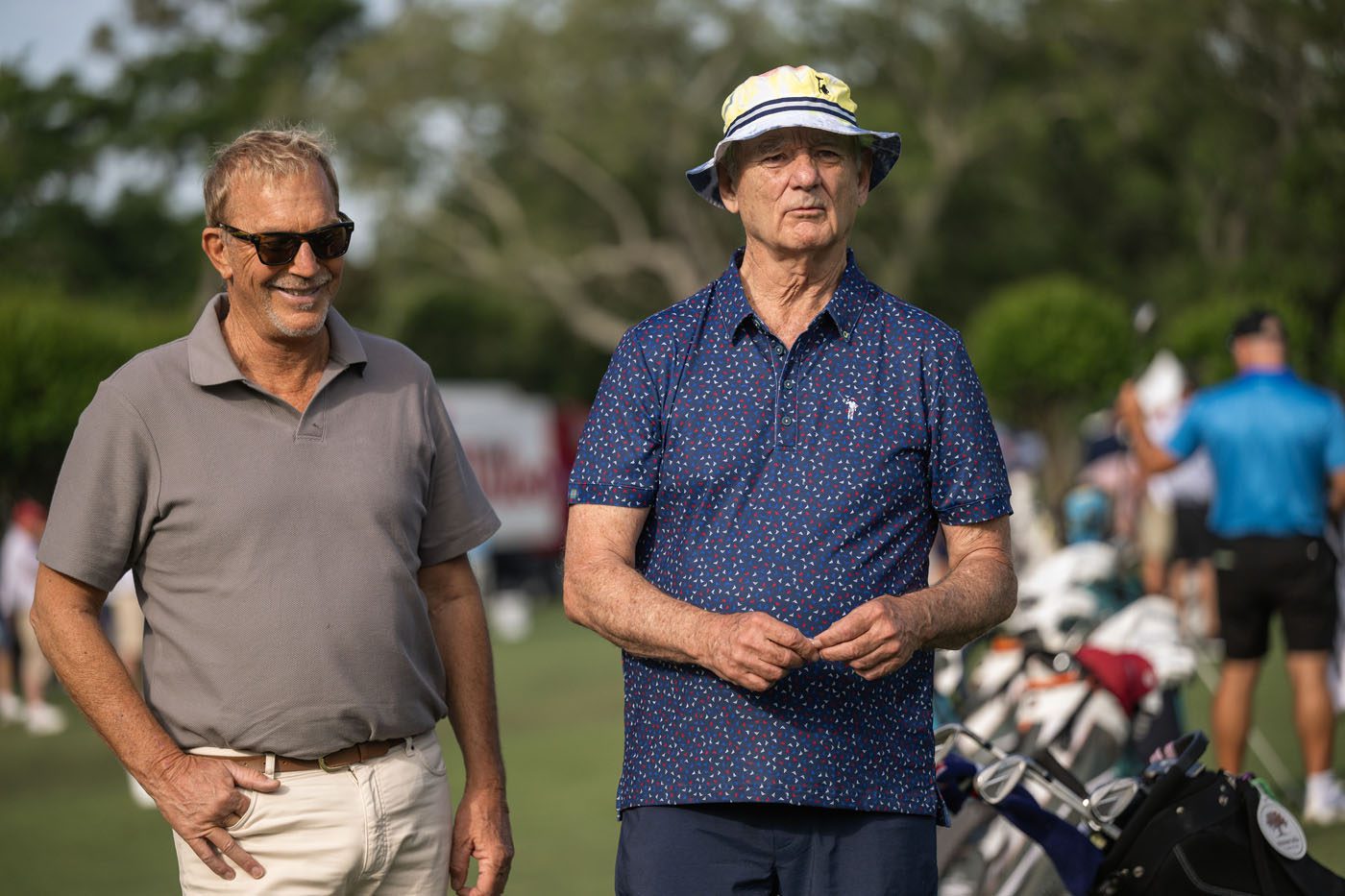 Two older men stand on a golf course, one wearing sunglasses and a polo shirt, the other in a patterned shirt and bucket hat. Golf bags and people are visible in the background.