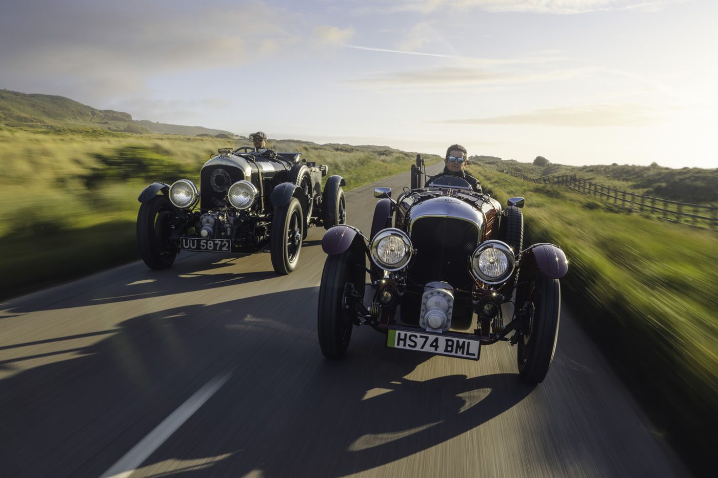 Two vintage cars, including a Blower Jnr, with drivers travel on a scenic road through the countryside. A blurred background suggests motion on this Bentley & Hedley Studios production, set against clear skies above.