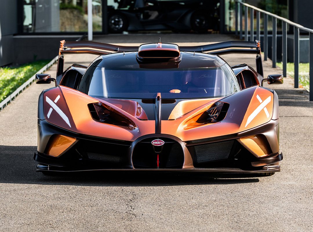 A front view of an orange and black $4.7Million Bugatti Bolide with copper carbon fiber accents and its doors open, parked on pavement near a modern building.