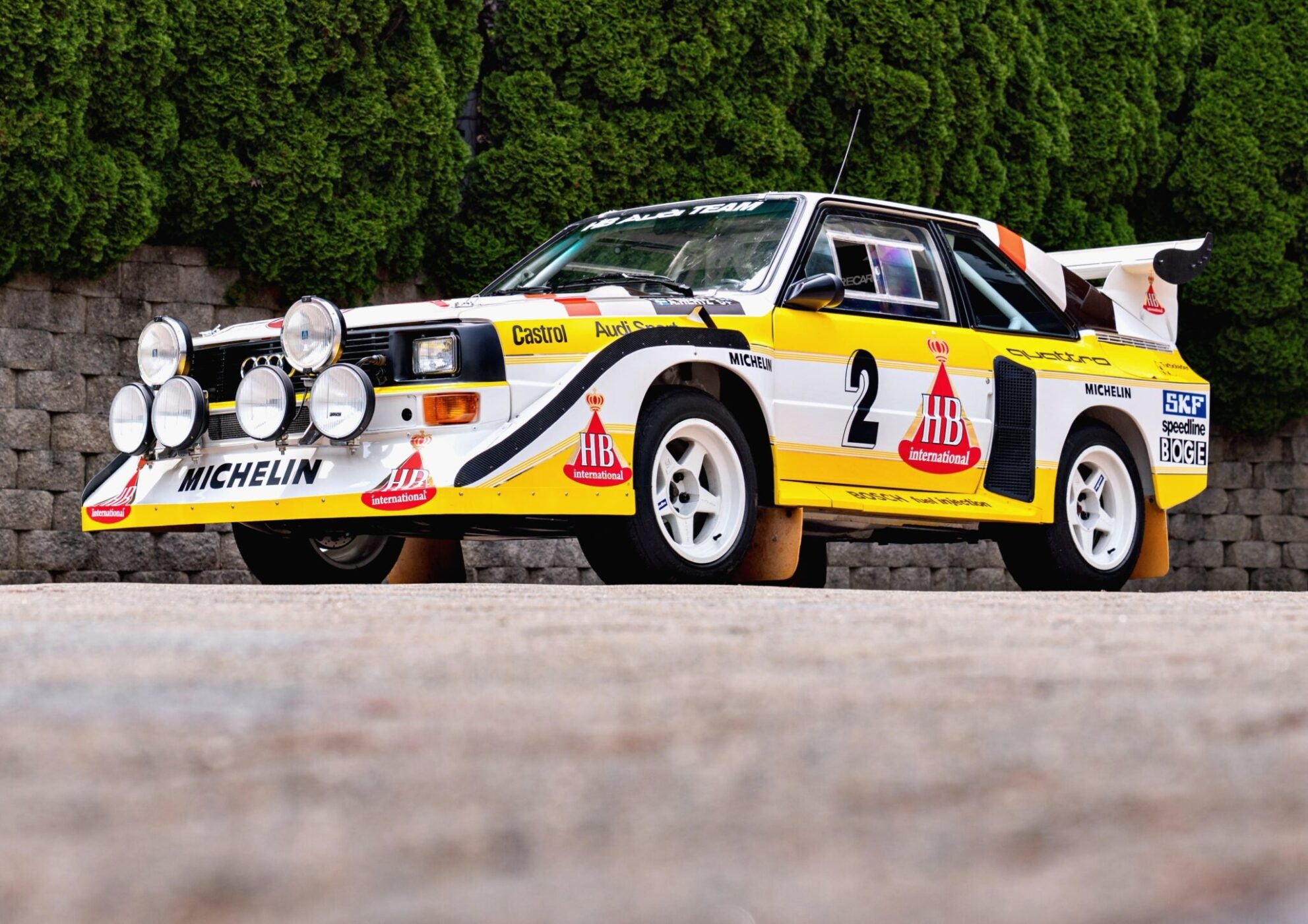 A white and yellow rally car with sponsor logos and multiple front-mounted spotlights is parked on a paved surface, highlighted as one of the 2025 Bonhams Monterey: 10 Must-See Cars Crossing the Block at The Quail.