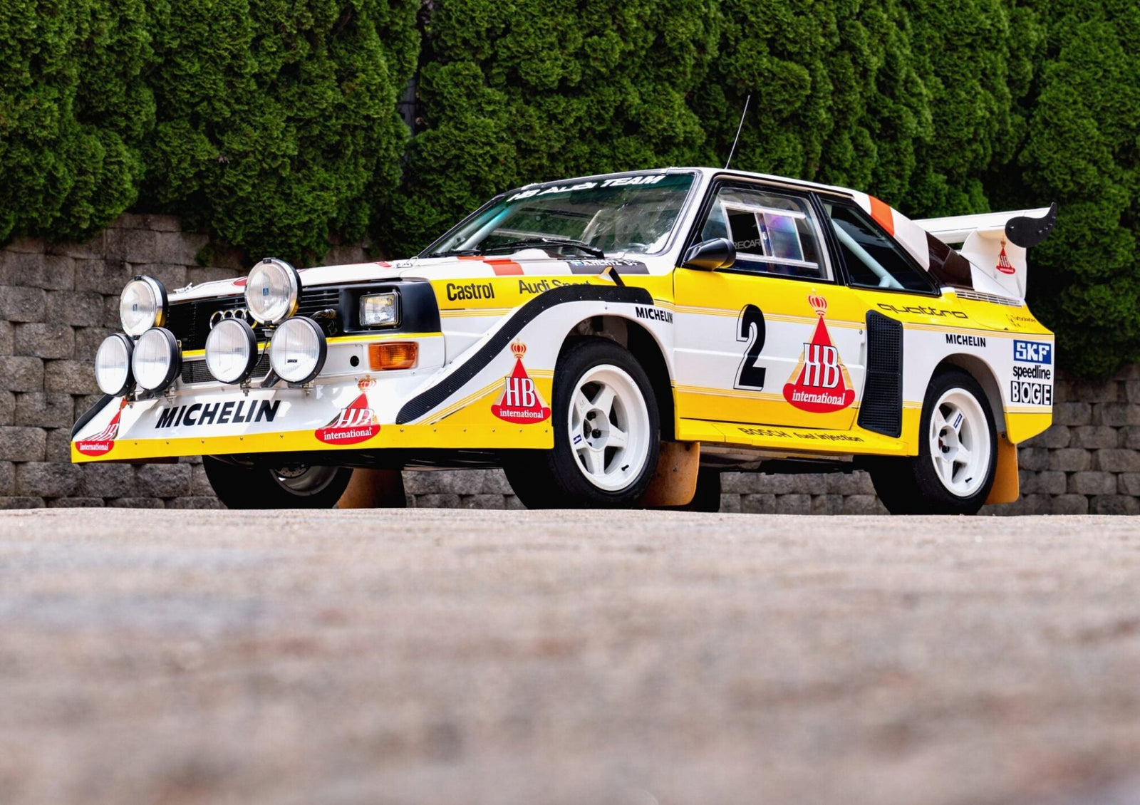 A white and yellow rally car with sponsor logos and multiple front-mounted spotlights is parked on a paved surface, highlighted as one of the 2025 Bonhams Monterey: 10 Must-See Cars Crossing the Block at The Quail.