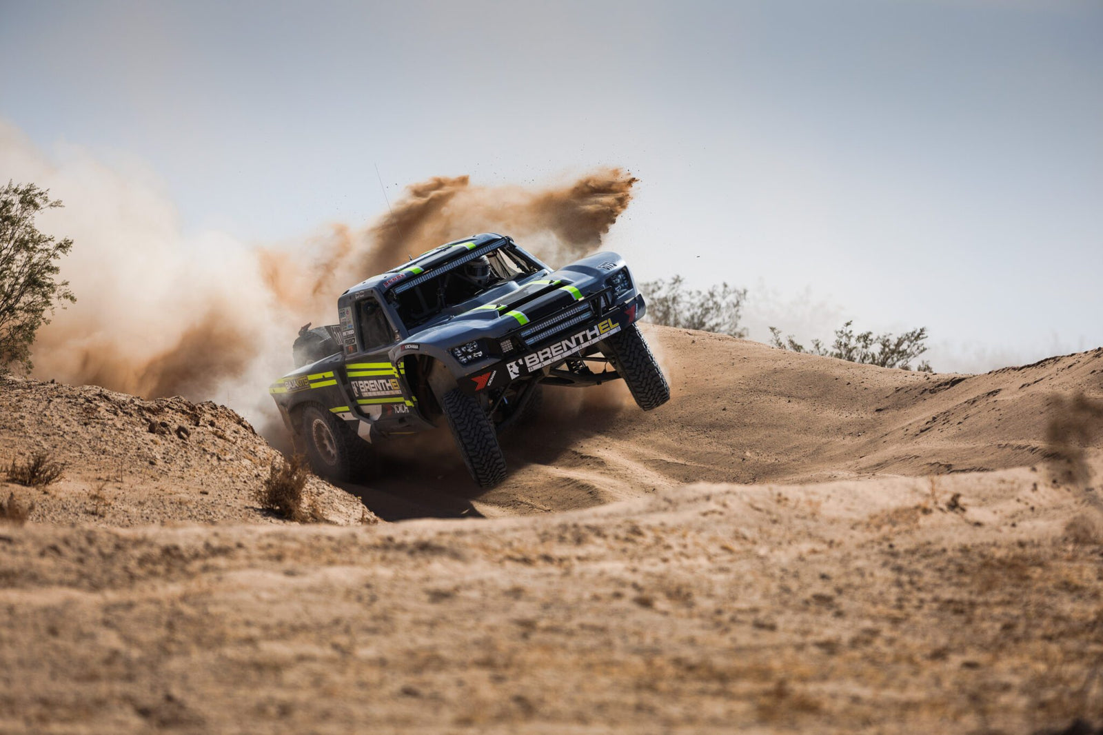 Off-road racing truck from Brenthel Industries kicks up dust while driving over a sandy dirt mound in a desert landscape, capturing the thrill of an Off-Road Adventure under clear skies.