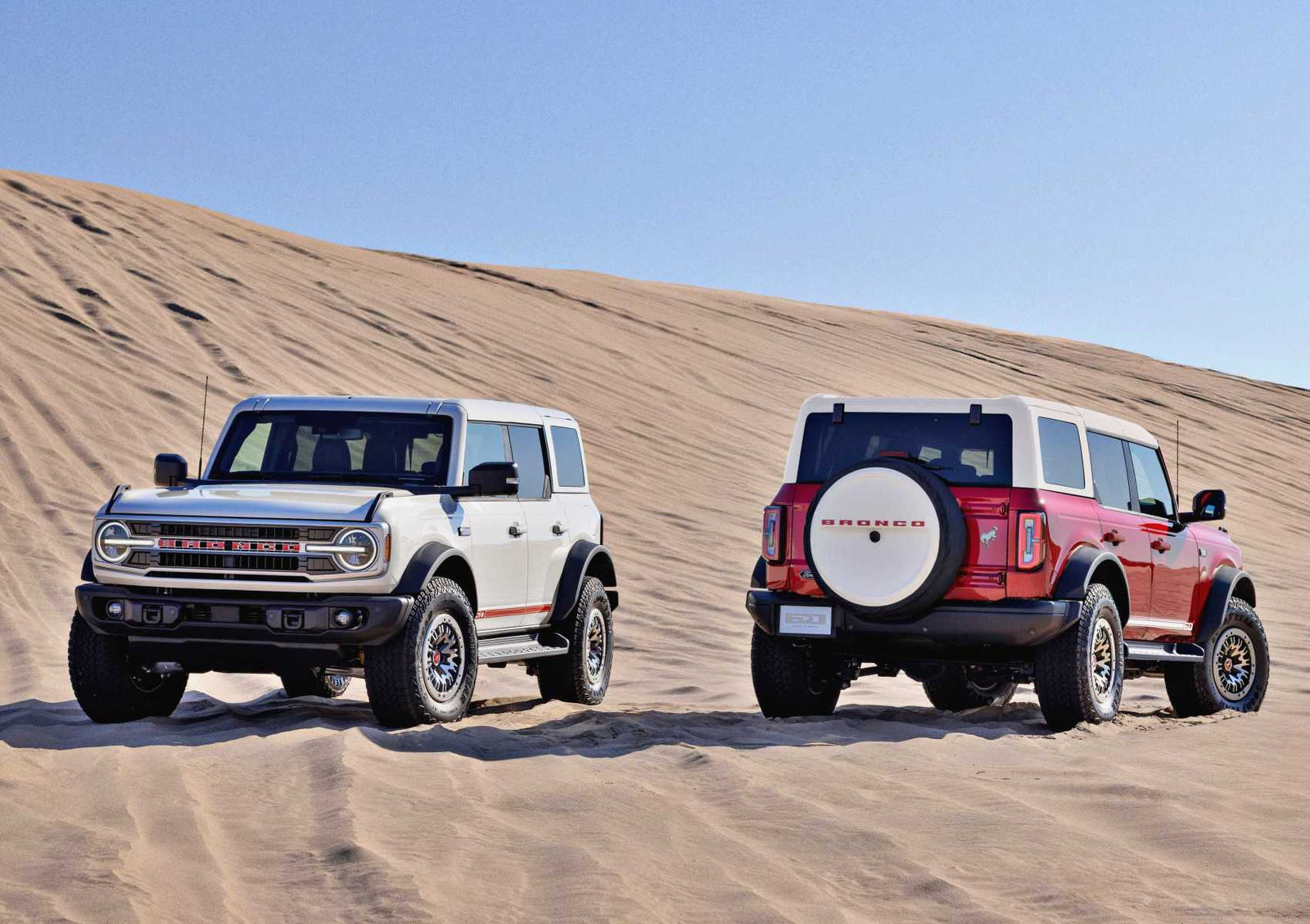 Two Ford Bronco SUVs, including a 2026 Ford Heritage Edition, are parked on sand dunes under a clear blue sky—one facing forward and the other backward.