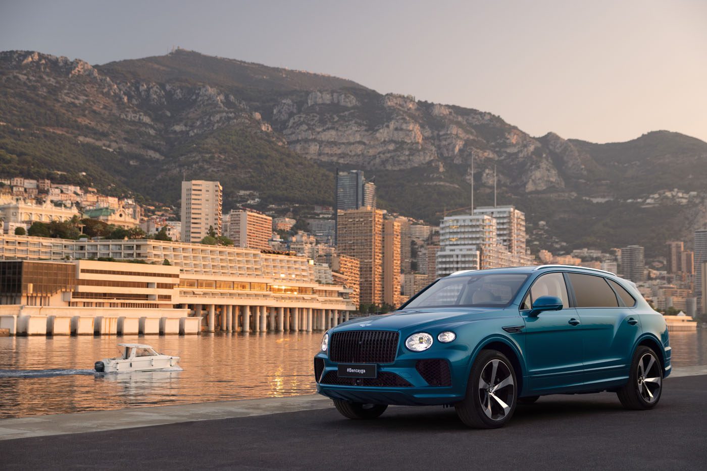 A blue Bentley Bentayga from the EWB Riviera Collection is parked by a waterfront with a city skyline and mountains in the background. A white boat glides through the water, creating a picturesque scene reminiscent of Brussels' finest views.
