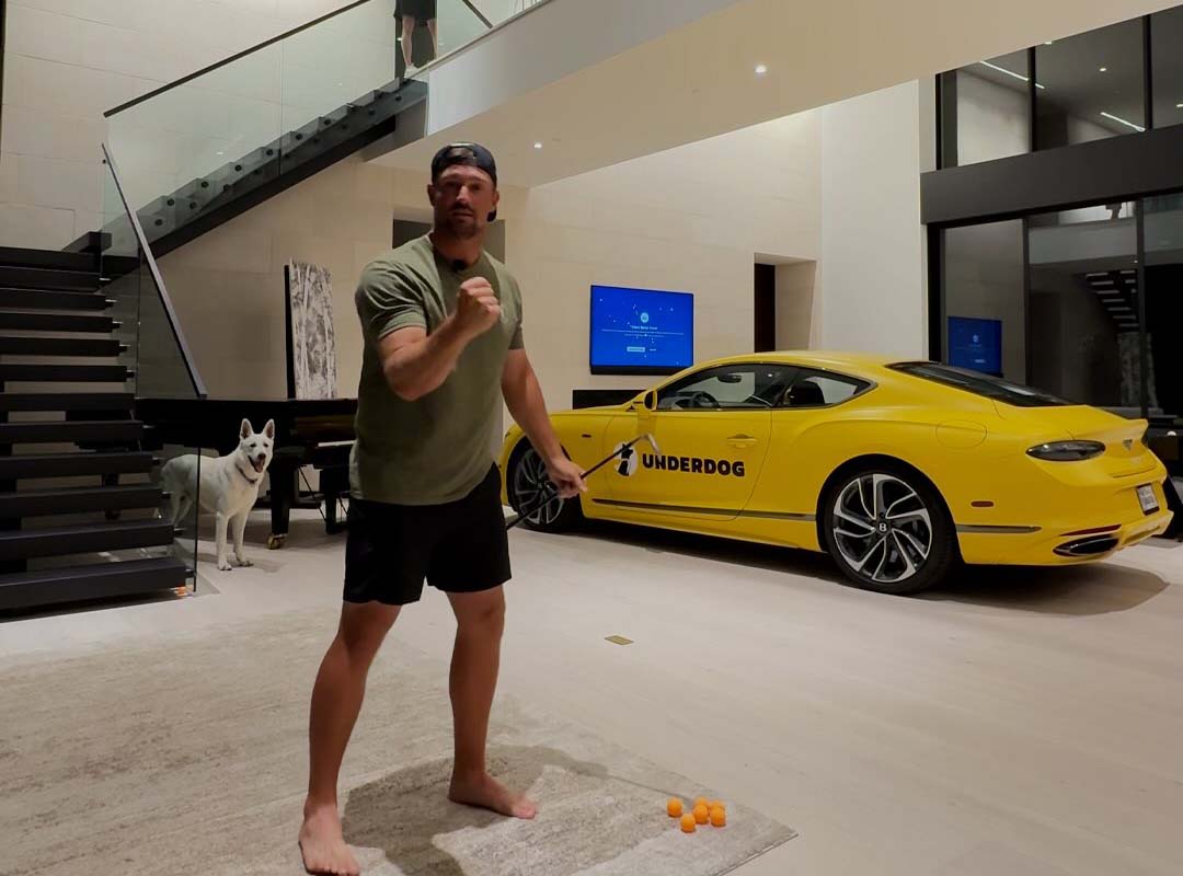 A man stands barefoot indoors in a ready stance for a Trick Shot Challenge, with a white dog nearby and a yellow Bentley Continental labeled “UNDERDOG” parked behind him.