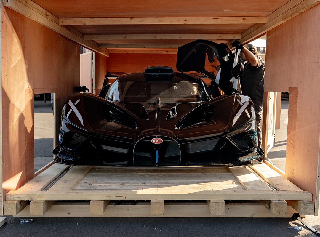 A Bugatti Bolide, valued at $4.7M, is being carefully secured inside a wooden crate, its door open while Manny Khoshbin adjusts it with precision.
