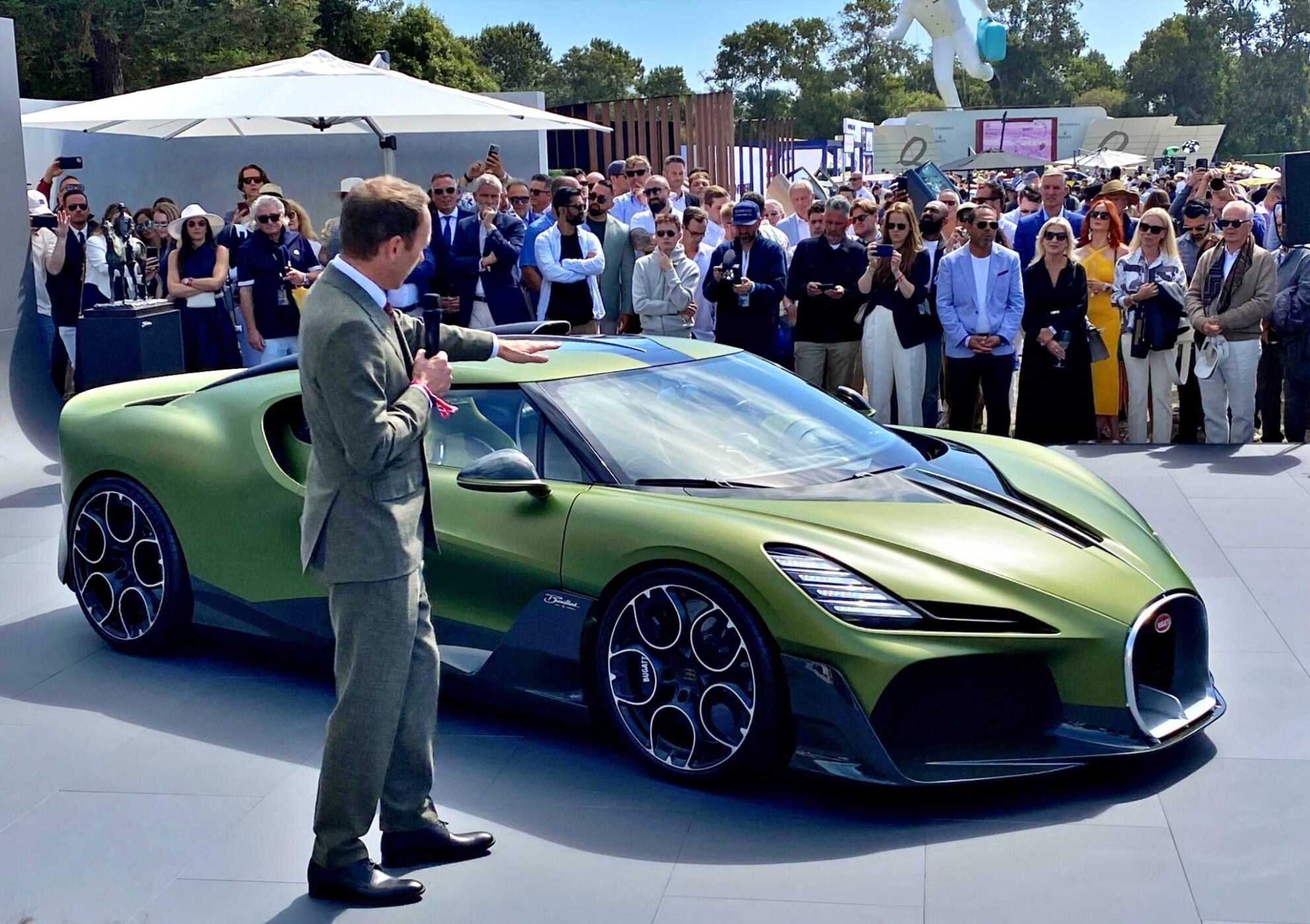A man in a suit presents a green Bugatti sports car to a crowd at an outdoor event on a sunny day.