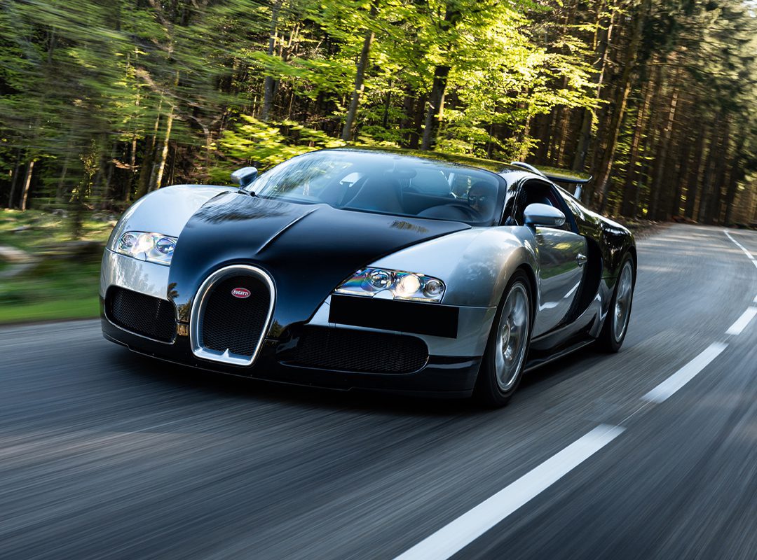 A black and silver Bugatti Veyron, featuring the advanced Chassis 5.0, speeds along a winding road through a lush, forested landscape.