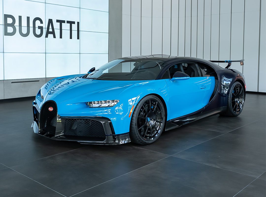 A blue and black Bugatti Chiron Pur Sport in striking French Racing Blue is on display in a modern showroom with large “BUGATTI” signage in the background, ready and available for sale.