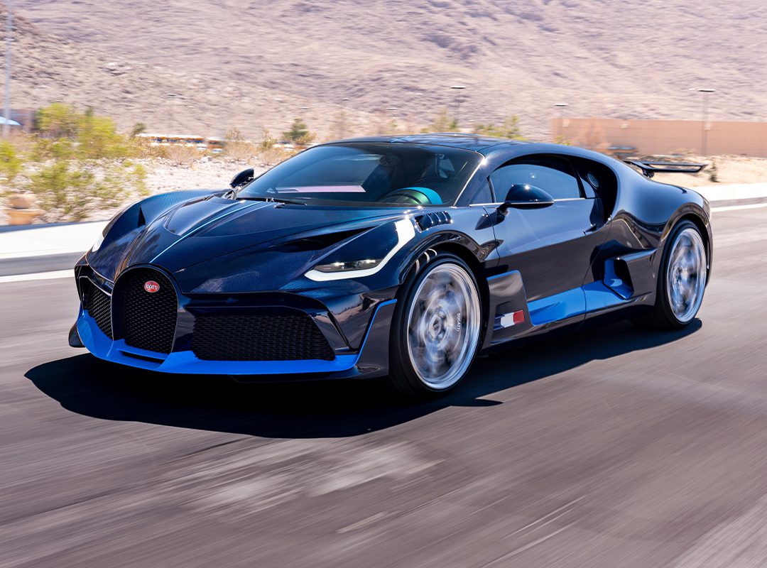 A blue carbon Bugatti Divo speeds along a paved road, set against a mountainous desert landscape, and is now for sale.