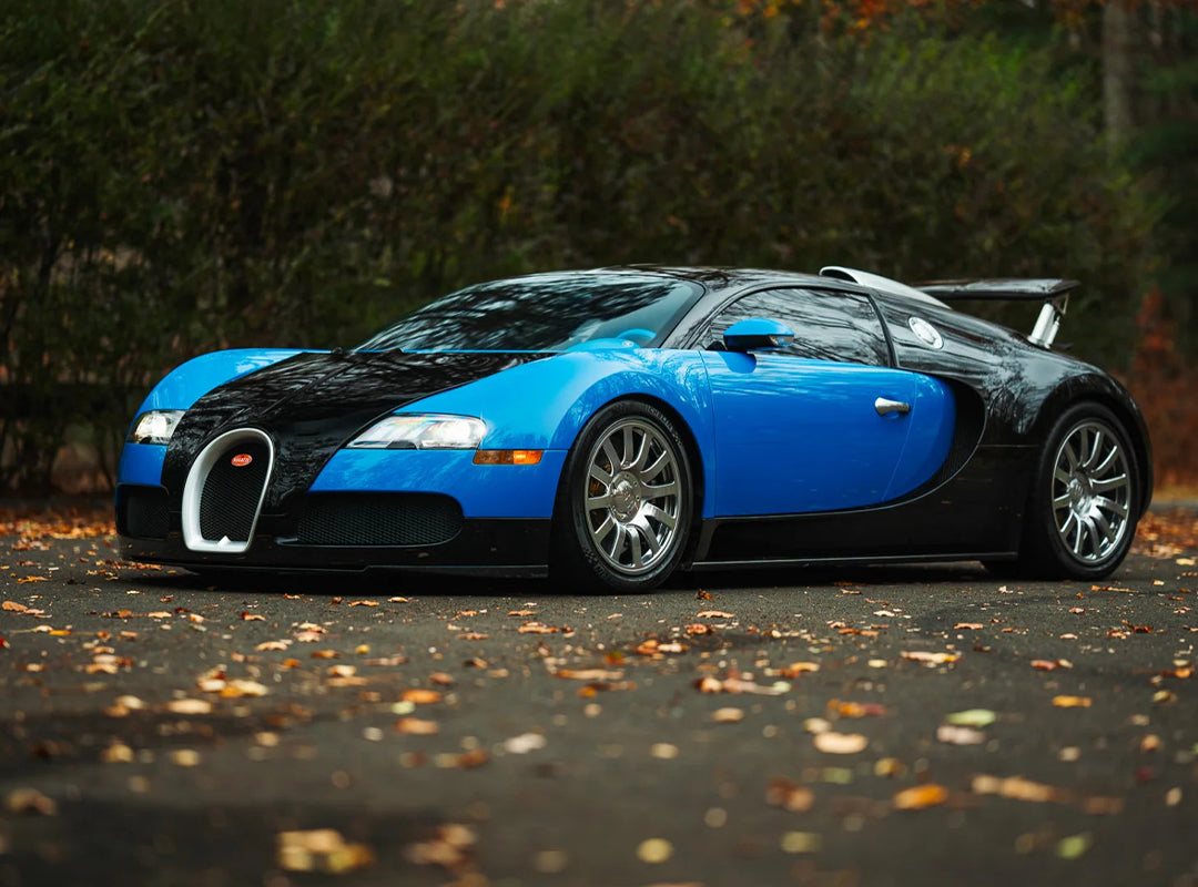 A blue and black Bugatti Veyron, one of the most expensive Bugatti hypercars of all time, is parked on a leaf-covered pavement with trees in the background.