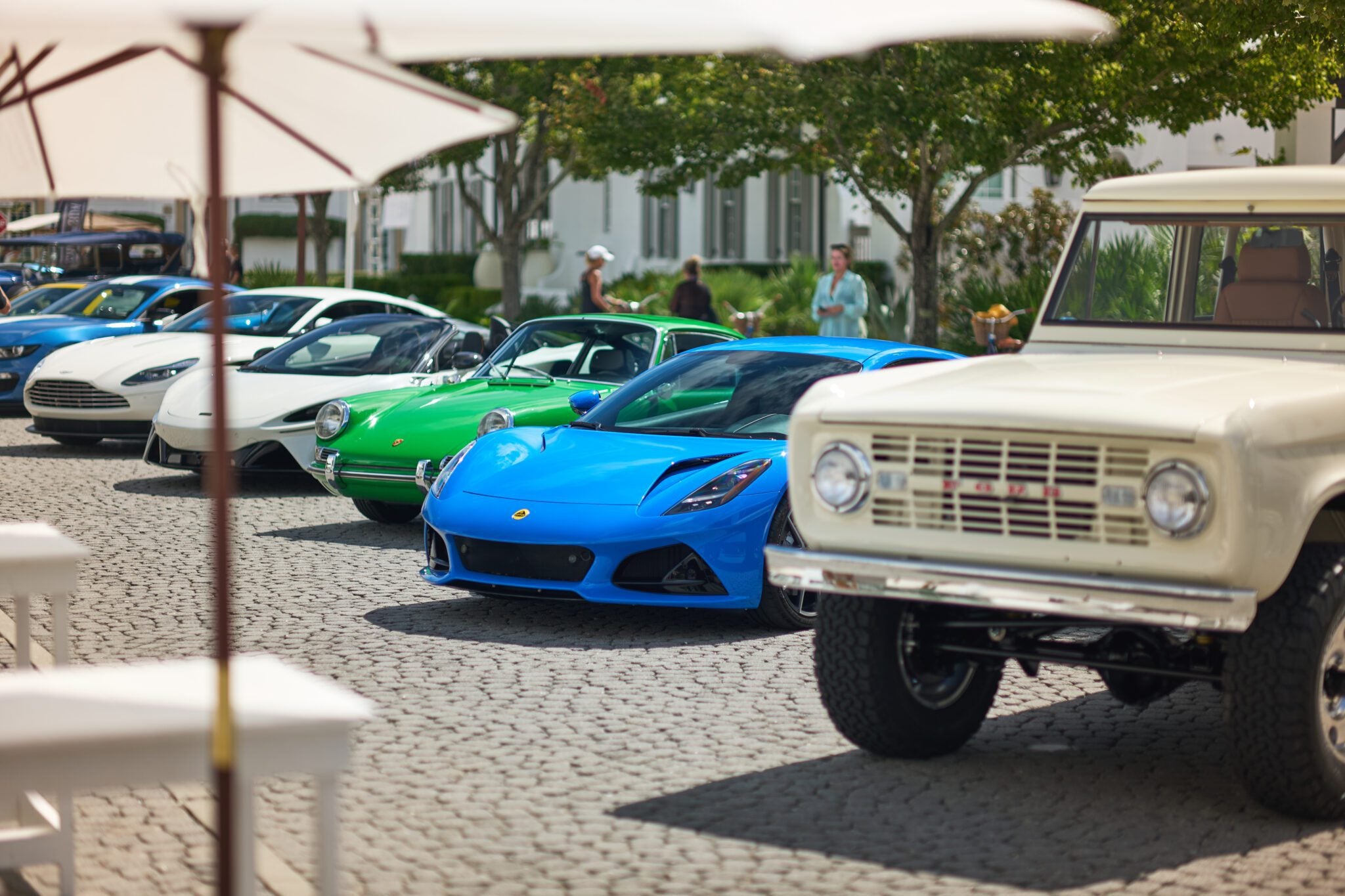 A row of classic and modern supercars, including a blue Lotus sports car, lines a cobblestone street at Cars of 30A, with people and trees in the background.