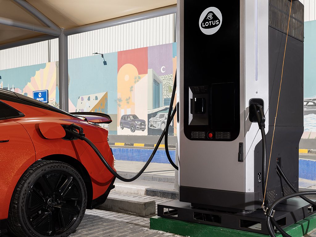 An orange electric car is charging at a Lotus-branded charging station under a canopy, with a colorful mural in the background.