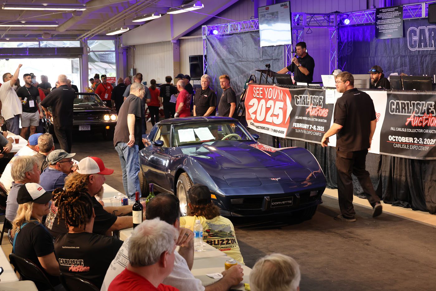 A blue Corvette is presented on stage at the Carlisle Auctions Spring Carlisle Auction, with attendees seated at tables and staff nearby. Signs display auction details and the date for the next Carlisle Auctions event in 2025.