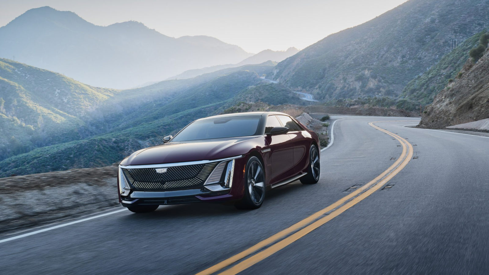 A maroon Cadillac sedan drives on a winding mountain road with green hills and blue sky in the background.