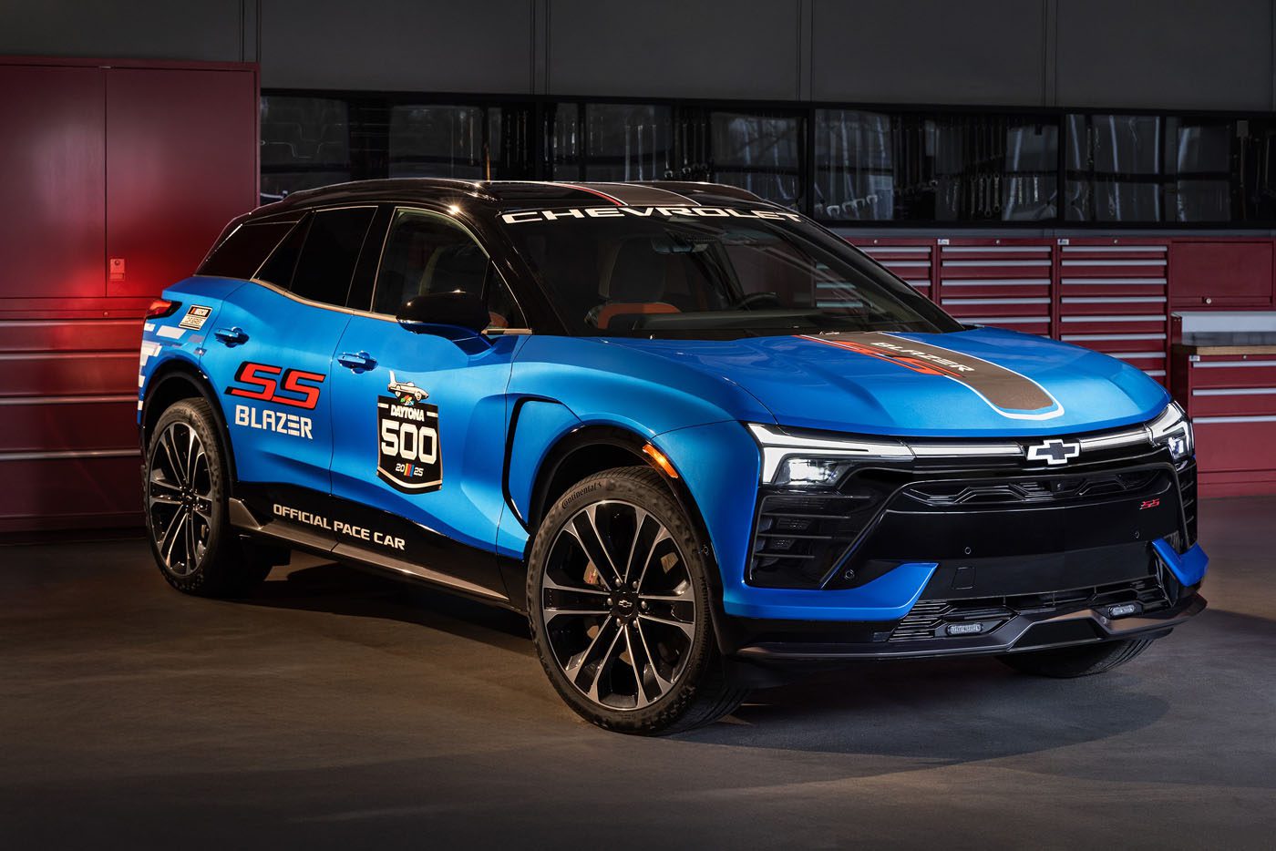 A blue Chevrolet Blazer SS with racing decals, echoing a sporty design, sits indoors near red tool cabinets—a vision reminiscent of a Pace Car ready for the Daytona 500.