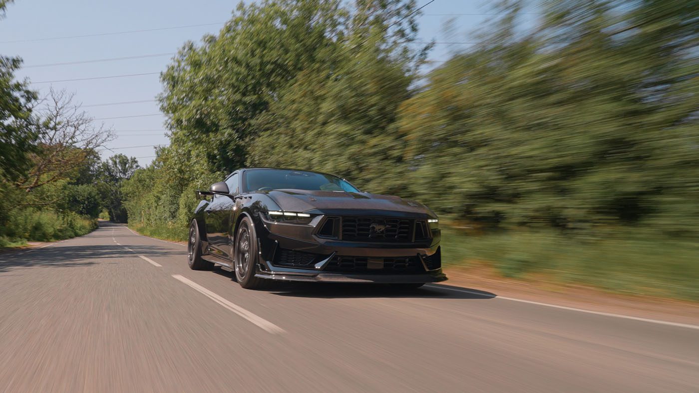 A Ford Mustang Dark Horse sports car speeds along a tree-lined, two-lane road under a clear sky, embodying the power and style of Clive Sutton’s renowned vehicles.
