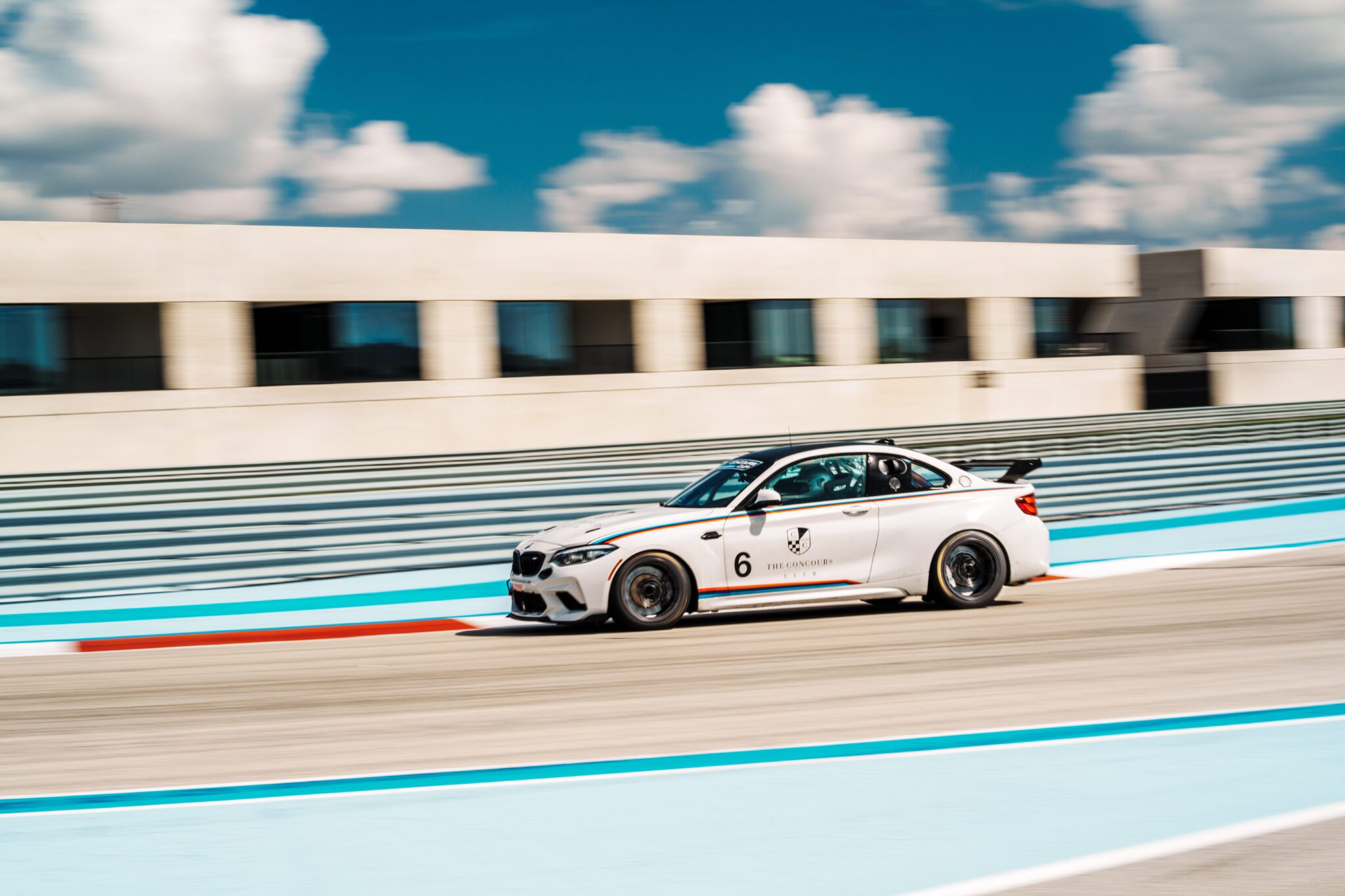 A sleek white race car races around the racetrack at Concours Club, its engine roaring in front of a modern building under a blue sky with scattered clouds.