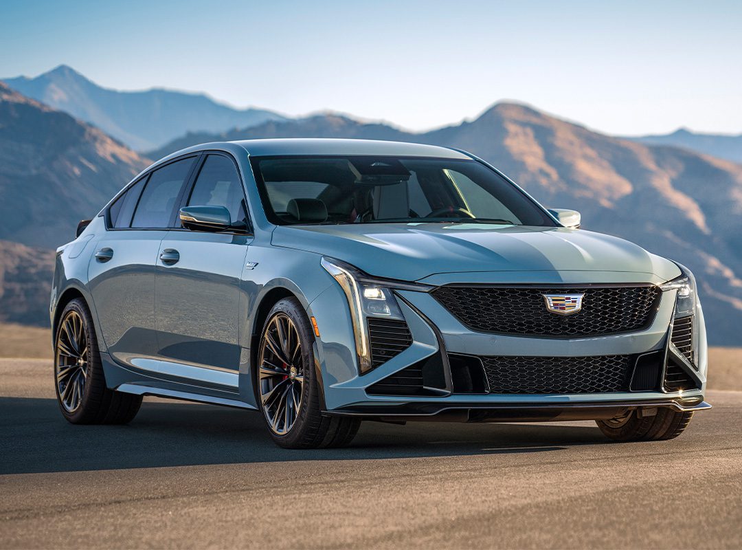 A silver Cadillac CT5-V Blackwing sedan with 668HP is parked on a paved surface with mountains in the background under clear skies.