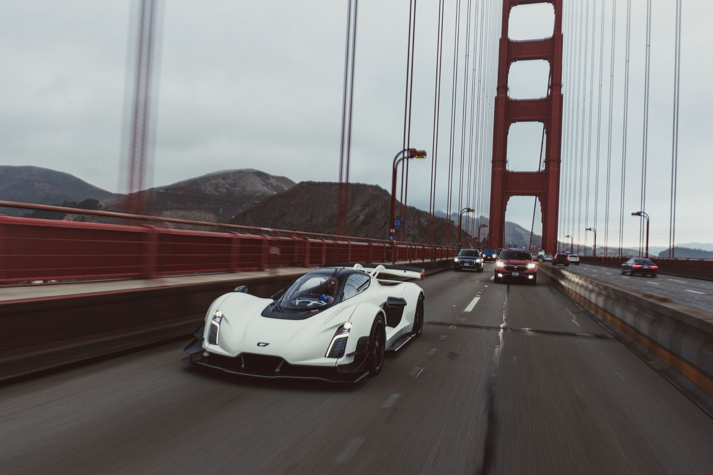A white sports car drives on a bridge alongside other vehicles under an overcast sky, with suspension cables and hills visible in the background.