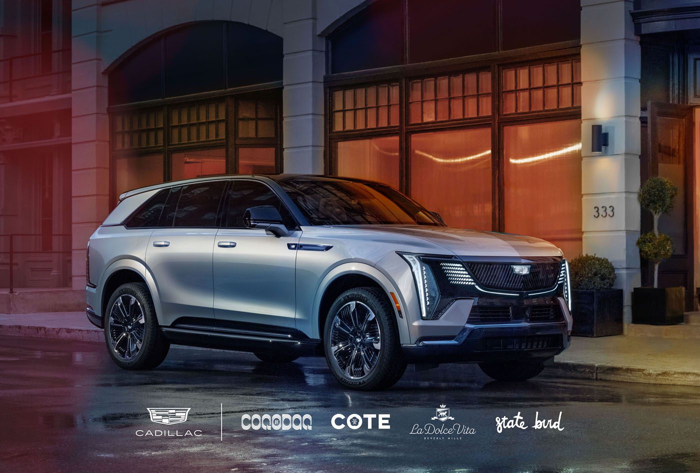 A silver SUV is parked on a city street in front of an illuminated building. Logos for Cadillac, Cote, La Dolce Vita, and others are displayed at the bottom of the image.