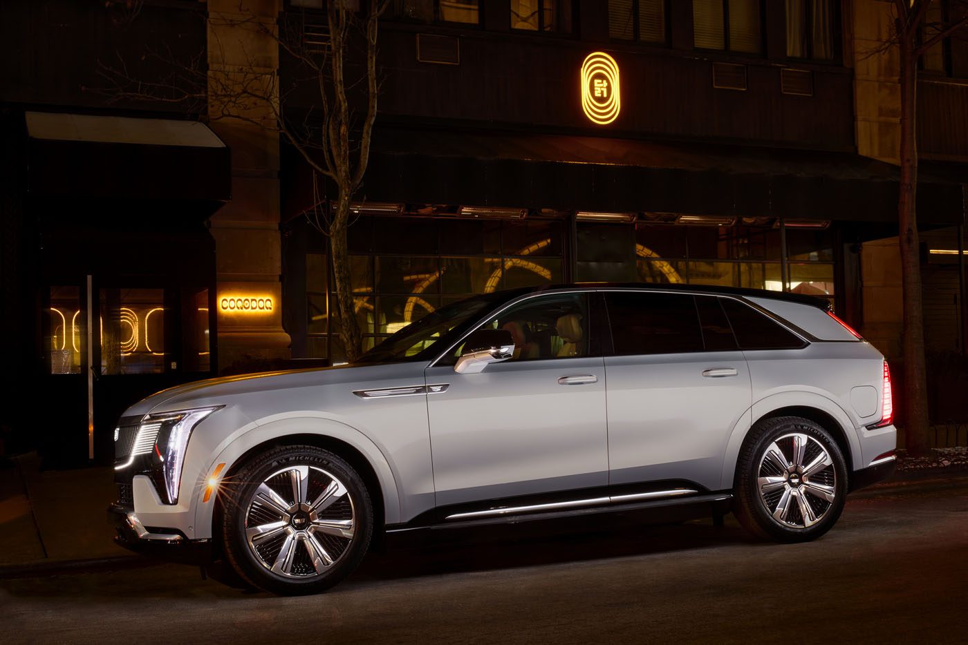 A silver Cadillac SUV, possibly the elegant Escalade IQ, sits parked on a dimly lit street with illuminated signage hinting at a nearby dining experience in the background.